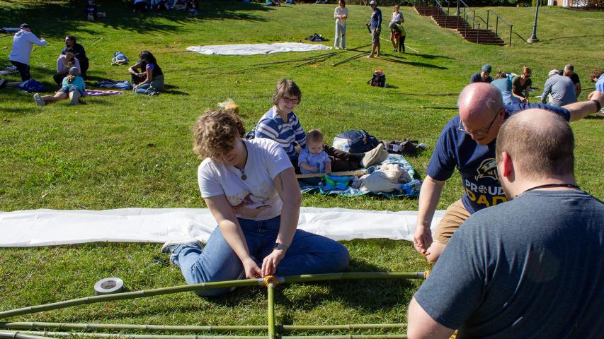Several people sit on the grass assembling a bamboo structure, while others relax on blankets in the background on a sunny day.