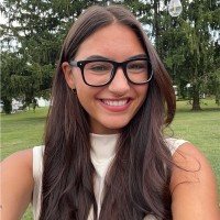 A woman with long brown hair and glasses smiles outdoors in front of green trees and grass.
