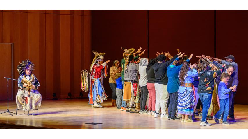 A group of people stands in a semi-circle on stage raising their arms, while two performers in traditional Native American attire and one seated musician are present.