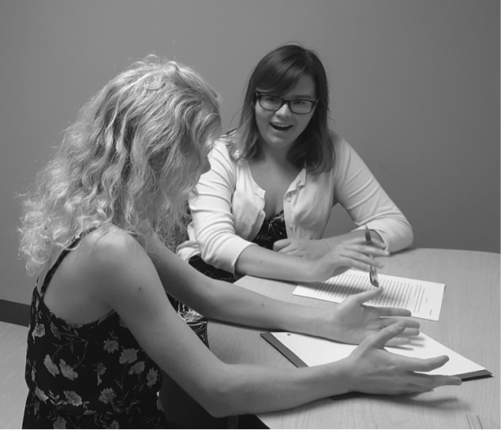 A tutor and student sit at a table discussing documents; the tutor gestures while speaking, the student holds a pen and listens attentively.
