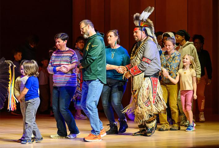 A group of people, including a man in traditional Native American attire, holding hands and dancing in a line on a stage.