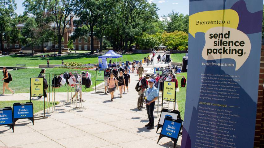 People walk through a campus exhibit displaying backpacks and informational signs for the "Send Silence Packing" mental health awareness event.