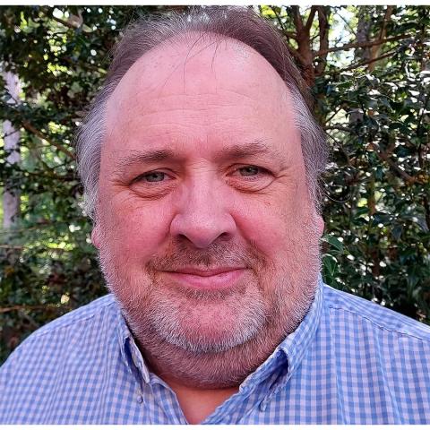 A middle-aged man with graying hair and a beard, wearing a blue checked shirt, stands outdoors in front of green foliage.