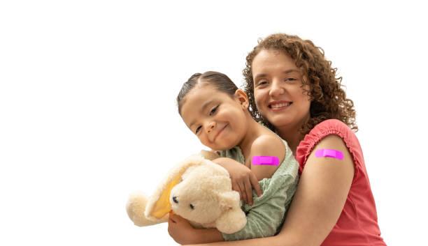 A woman and a child with pink bandages on their arms smile at the camera; the child is holding a teddy bear.