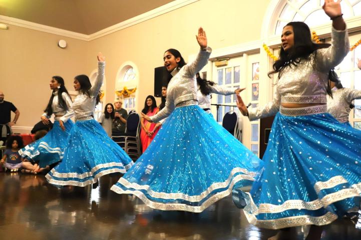 A group of women in silver tops and blue skirts perform a synchronized dance routine indoors, with onlookers seated and standing in the background.