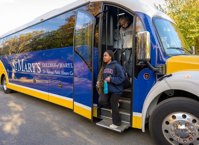 Two students step off a St. Mary's College of Maryland River Runner bus parked on a road, with one holding a water bottle and both carrying bags.