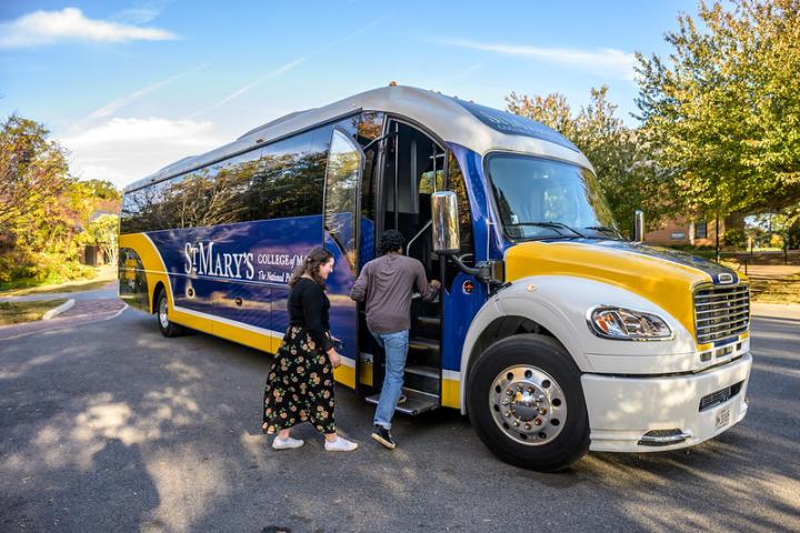 Two people are boarding a blue and yellow St. Mary's College of Maryland River Runner bus parked on a tree-lined street.