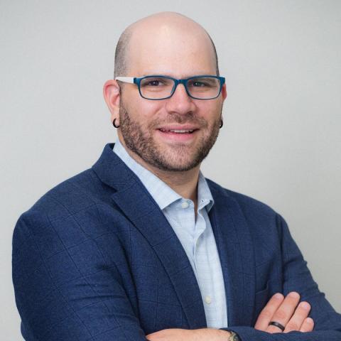 A man with a brown beard in a blue sport coat with pale blue shirt wearing glasses, looking at the camera with arms folded across chest.