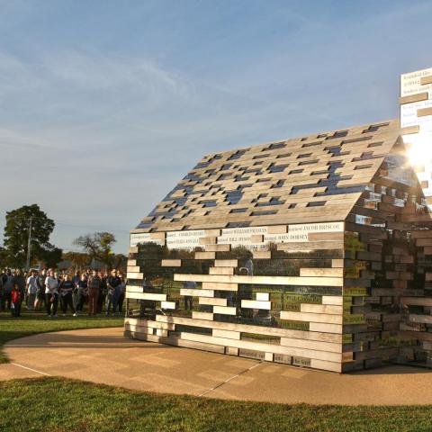 A group of people observe a large outdoor sculpture shaped like a house, constructed from rectangular reflective metal blocks, on a grassy field under a clear sky.