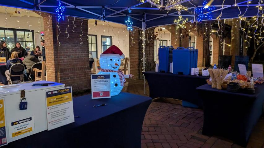 Outdoor event setup with tables under string lights, a snowman decoration, beverage dispensers, snacks, and people sitting in the background.
