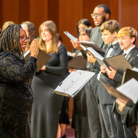 A choir conductor leads a group of singers holding sheet music during a performance on stage.