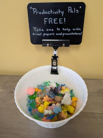 A white bowl filled with small colorful figurines sits on a table beneath a sign reading "'Productivity Pals' Free! Take one to help with final papers and presentations!.