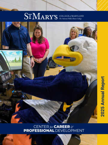 A person in an eagle mascot costume sits at a driving simulator while several people stand nearby, smiling, in a gymnasium setting.