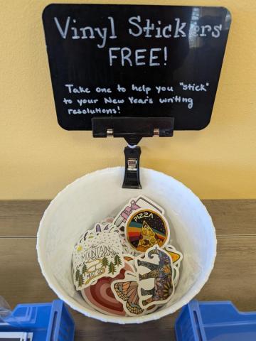 A white bowl holding assorted vinyl stickers sits on a wooden surface. Above it, a sign reads: "Vinyl Stickers FREE! Take one to help you 'stick' to your New Year's writing resolutions!.