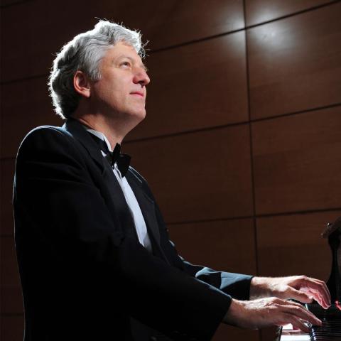 A man in a tuxedo plays a grand piano on stage, with wooden wall panels behind him and focused stage lighting.