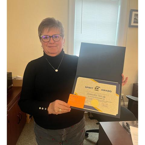 A person wearing glasses holds an open folder displaying a "Spirit Award" certificate and an orange card in an office setting.