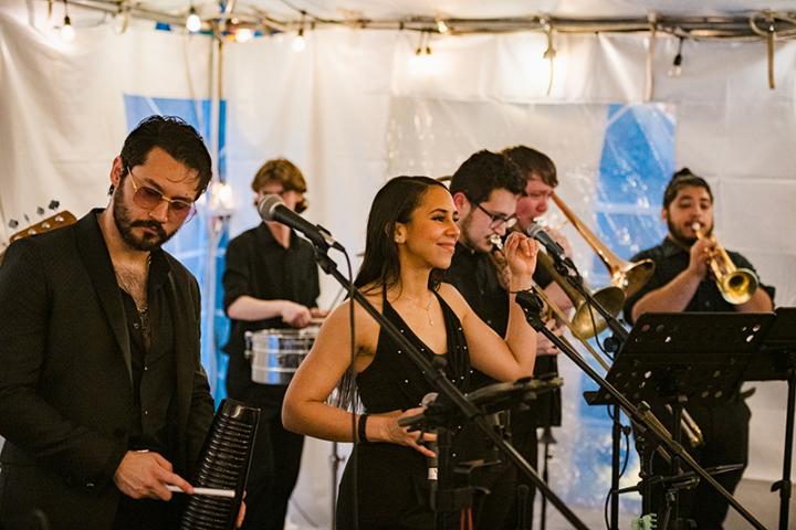 A band performs indoors; a woman sings into a microphone while men play percussion, trombone, and trumpet instruments.