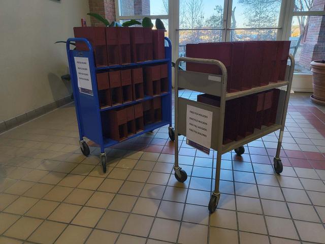 Two carts with maroon file folders stand in a tiled hallway near glass doors; signs on the carts indicate they hold files for free pickup.