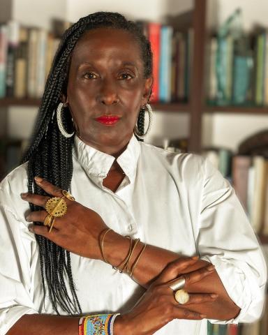 A person with long braided hair, wearing a white shirt and multiple rings, bracelets, and earrings, stands with arms crossed in front of a bookshelf.