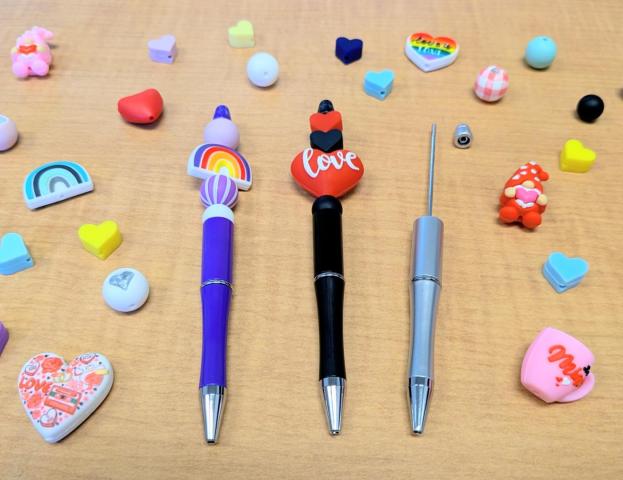Three decorative beaded pens with heart and rainbow designs are placed on a wooden surface surrounded by colorful beads in heart and other whimsical Valentine's shapes.