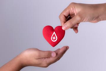 One hand holds a red heart with a medical symbol, while another hand reaches toward it, symbolizing blood donation or healthcare support.