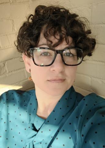 Person with short curly hair and large glasses wearing a teal polka dot blouse, sitting in front of a light-colored brick wall.