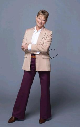 A person stands confidently with arms crossed, wearing a beige blazer, white shirt, purple trousers, and brown shoes, against a plain gray background.