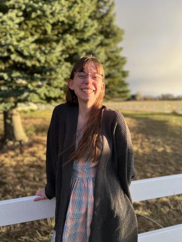 A person with long brown hair and glasses, wearing a cardigan and dress, stands outdoors by a white fence with trees and a field in the background.