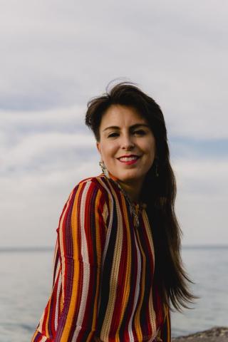 Woman with long dark hair wearing a red and yellow striped top stands outdoors near water, smiling at the camera under a cloudy sky.