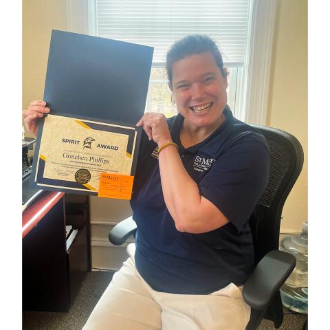 A woman sitting in an office chair smiles and holds up a certificate labeled "Spirit Award" with the name Gretchen Phillips visible.