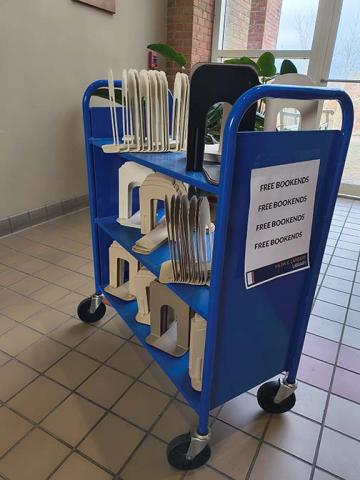 A blue cart with a sign offering free bookends, holding multiple white and black metal bookends, stands in a tiled hallway near windows and plants.