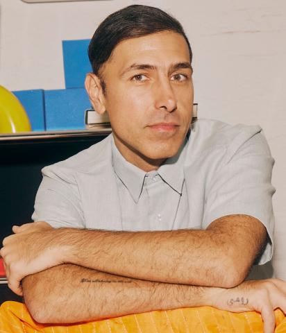A person with short dark hair and a light grey shirt sits with arms crossed on a table covered by an orange cloth, with colorful objects in the background.