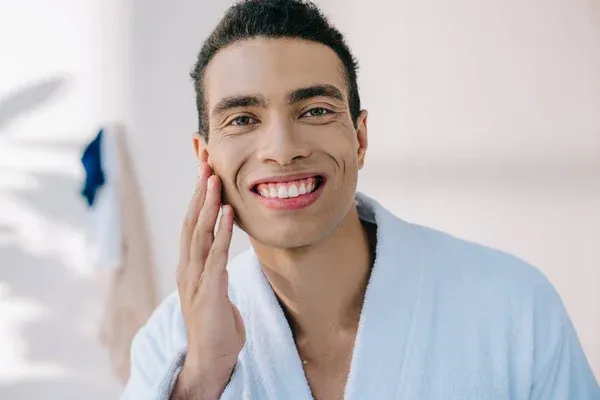 A man in a white bathrobe smiles and touches his cheek, standing indoors with towels hanging in the background.