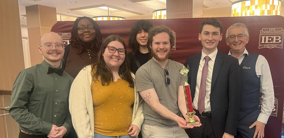 Seven people stand indoors; one holds a trophy. They pose in front of a maroon backdrop with a logo, and all are smiling at the camera.