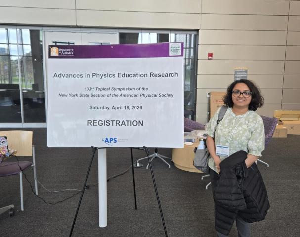 A person stands next to a sign for the "Advances in Physics Education Research" symposium, showing event details and a registration area inside a modern building.