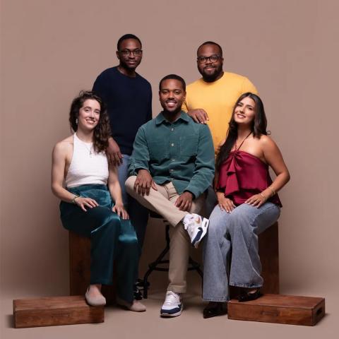 Five people pose together against a beige background, with two seated in front and three standing behind them, all smiling at the camera.