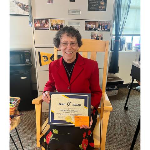 A woman in a red blazer sits in an office and smiles while holding a framed Spirit Award certificate and an orange gift card.