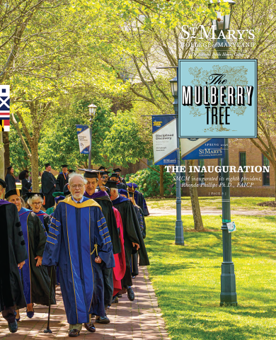 A group of people in academic regalia walk outdoors under trees. Banners and signs for St. Mary’s College of Maryland are visible.