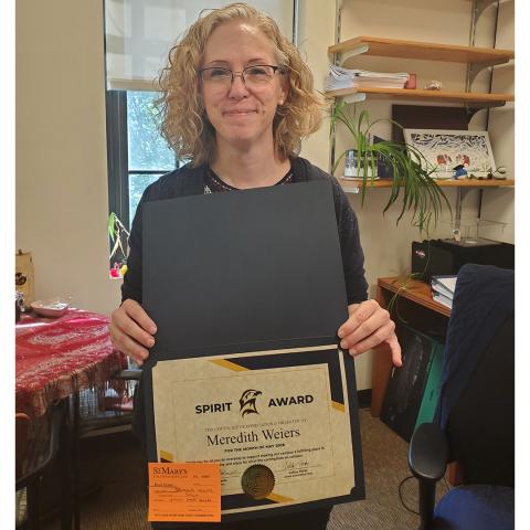 A woman with curly hair and glasses holds an open folder displaying a "Spirit Award" certificate and a small orange card in an office setting.