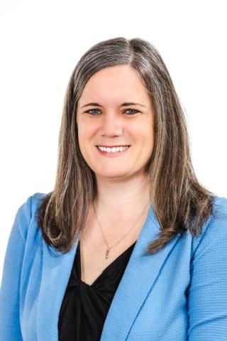 Woman with long brown and gray hair wearing a light blue blazer and black top, smiling, posed in front of a white background.
