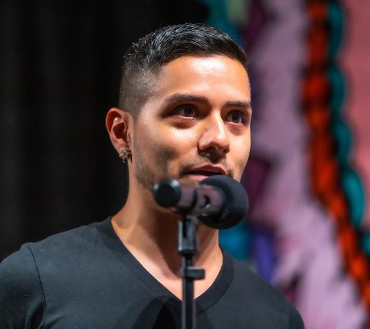 A person with short dark hair and earrings speaks into a microphone, wearing a black shirt. The colorful, blurred background suggests a lively setting.