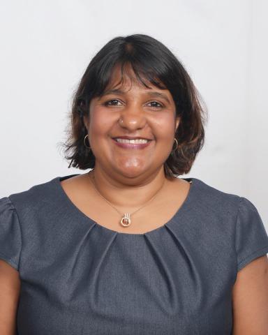 Woman with short brown hair, wearing a gray blouse, gold hoop earrings, and a gold necklace, smiles in front of a plain white background.
