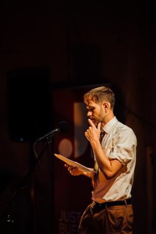 Joe Hall stands at a microphone, holding papers and touching his face thoughtfully, under warm stage lighting.
