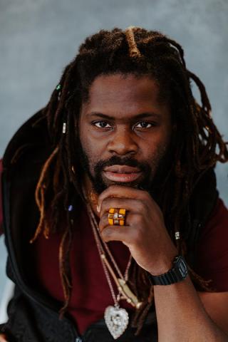 Man with long dreadlocks and a beard poses with his hand on his chin, wearing a maroon shirt, jewelry, a ring, and a black watch against a gray background.