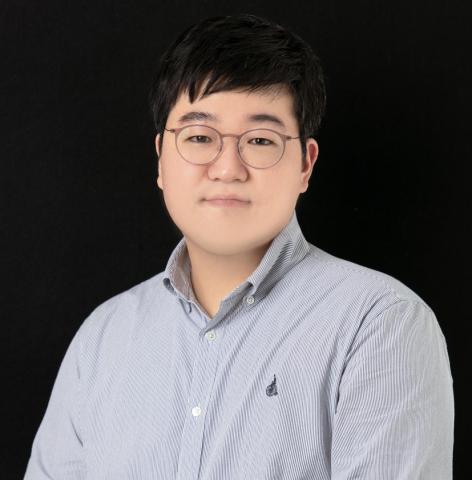 A person with short black hair and glasses wearing a light striped button-up shirt poses in front of a plain black background.