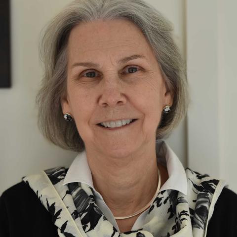 Older woman with gray hair smiles at the camera, wearing a white collared shirt, black patterned scarf, and earrings, standing indoors against a light-colored background.