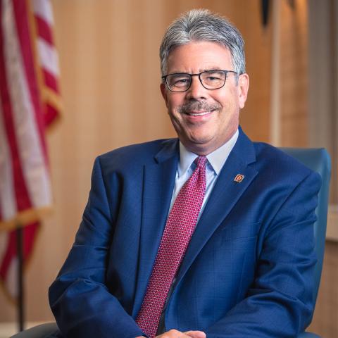 Man in a blue suit and red tie sits and smiles in an office setting with an American flag blurred in the background.