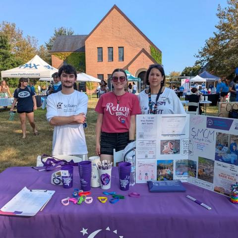 Three people stand behind a Relay for Life information table outdoors at a campus event, with brochures, cups, and bracelets displayed in front of them and a brick building in the background.