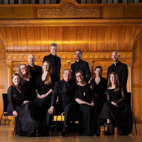 A group of eleven adults dressed in formal black attire pose together indoors in front of ornate wooden paneling.