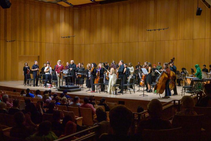 A symphony orchestra stands on stage with instruments, facing an applauding audience in a concert hall with wooden walls.